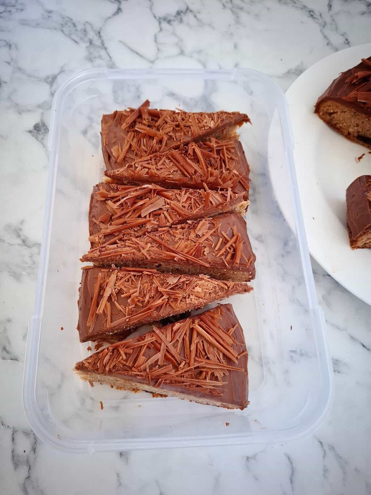 Six pieces of banana cake with chocolate icing in a rectangular plastic tub on a marbled bench top. The corner of a round, white plate with the rest of the cake next to it.