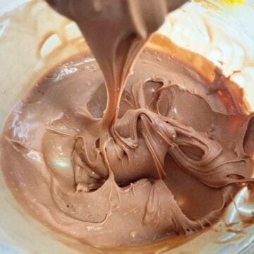 Chocolate ganache has cooled down to a thick but spreadable consistency good for icing a cake. The ganache is in a glass bowl and a spoon holding some up above it with some ganache slowly flowing down into the bowl.