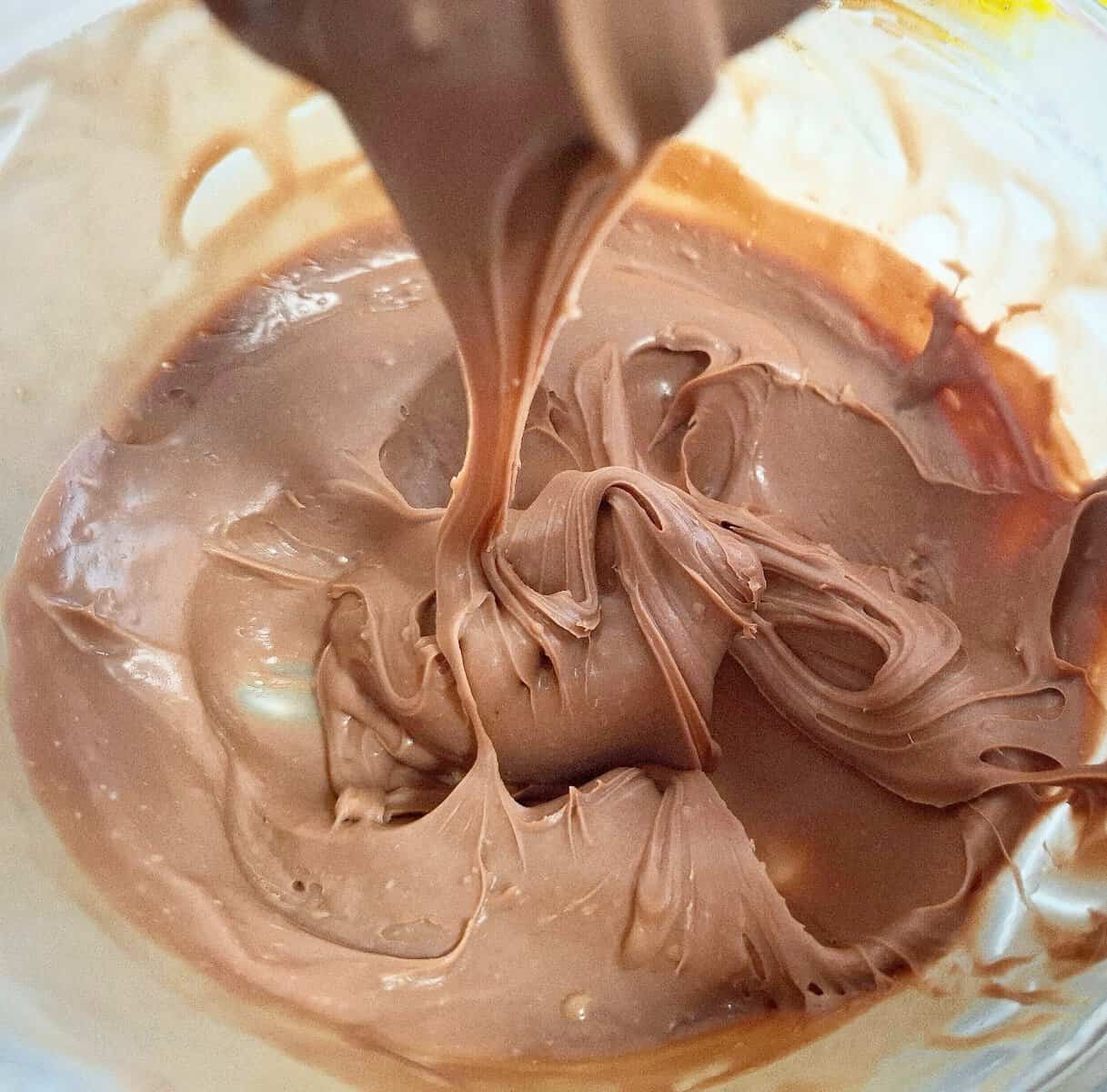 Chocolate ganache has cooled down to a thick but spreadable consistency good for icing a cake. The ganache is in a glass bowl and a spoon holding some up above it with some ganache slowly flowing down into the bowl.