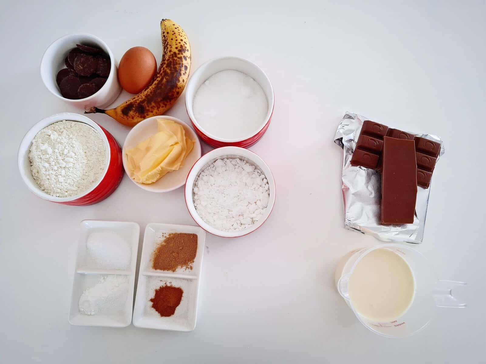 All ingredients for banana cake with chocolate icing in ramekins on a white table.