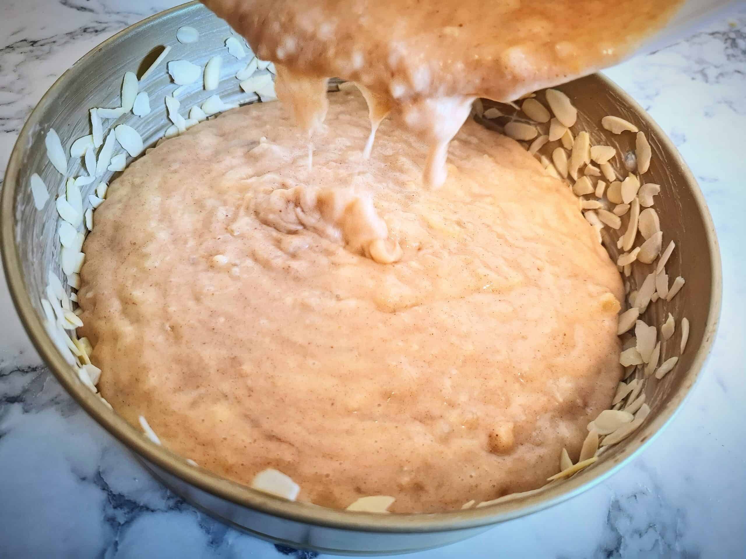 Pouring apple and almond cake batter into the prepared cake tin on a marbled bench top