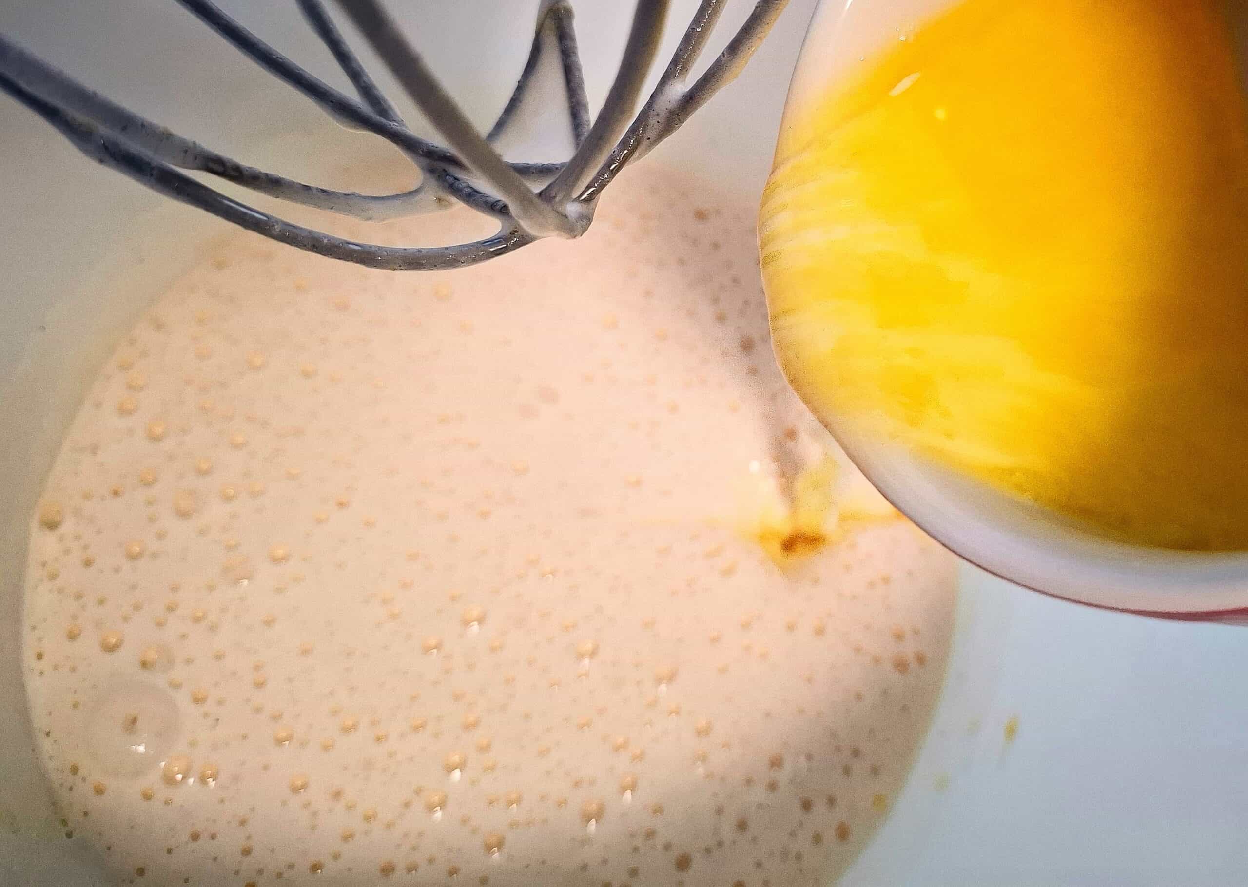 Melted butter and milk mixture being added from a white ramekin to the egg and sugar mixture in a white kitchen mixer bowl with a whisk above it.