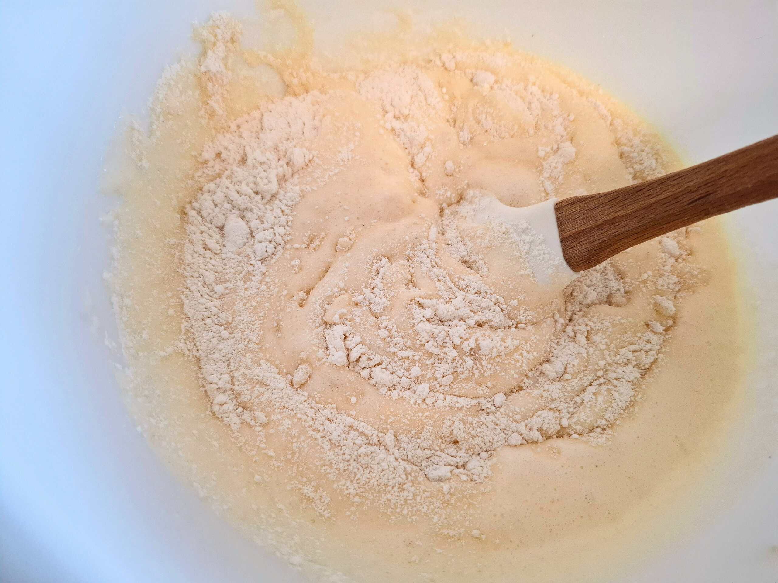 Flour mixture being gently folded into the cake batter in a white bowl with a wooden spoon.