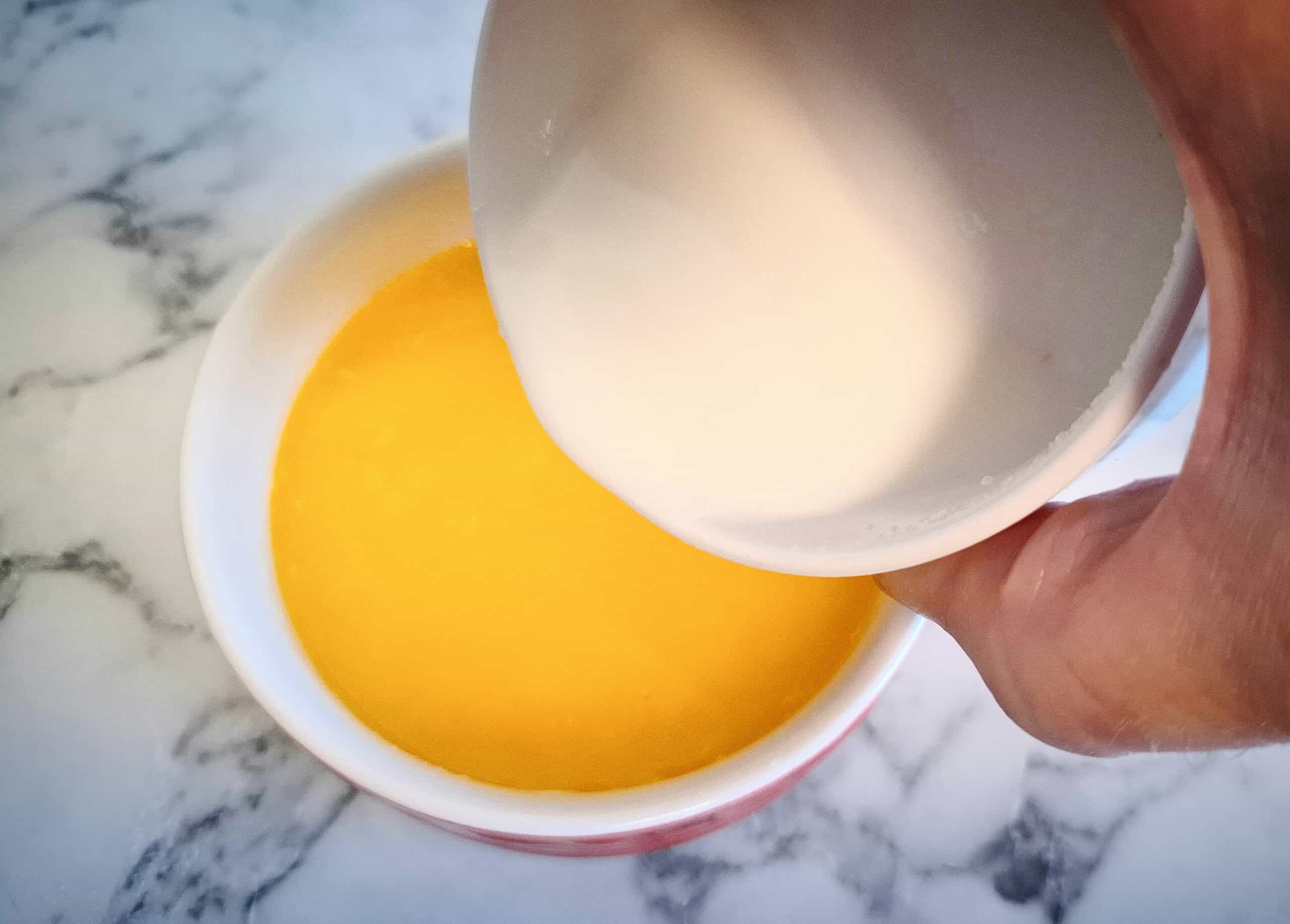 Melted butter in a white ramekin on a marbled bench top. A hand pouring milk into the butter from another white ramekin.