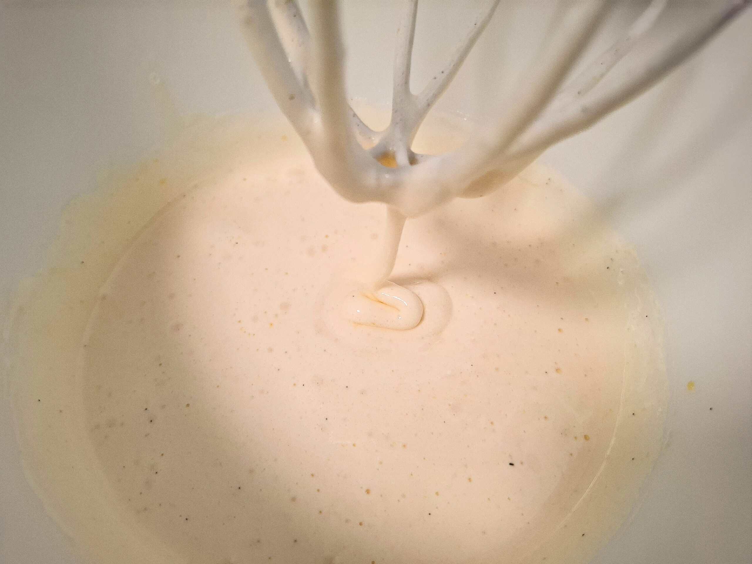Eggs, sugar and vanilla has been mixed white and fluffy in a white kitchen mixer bowl. A whisk with some egg mixture drizzling down into the bowl.