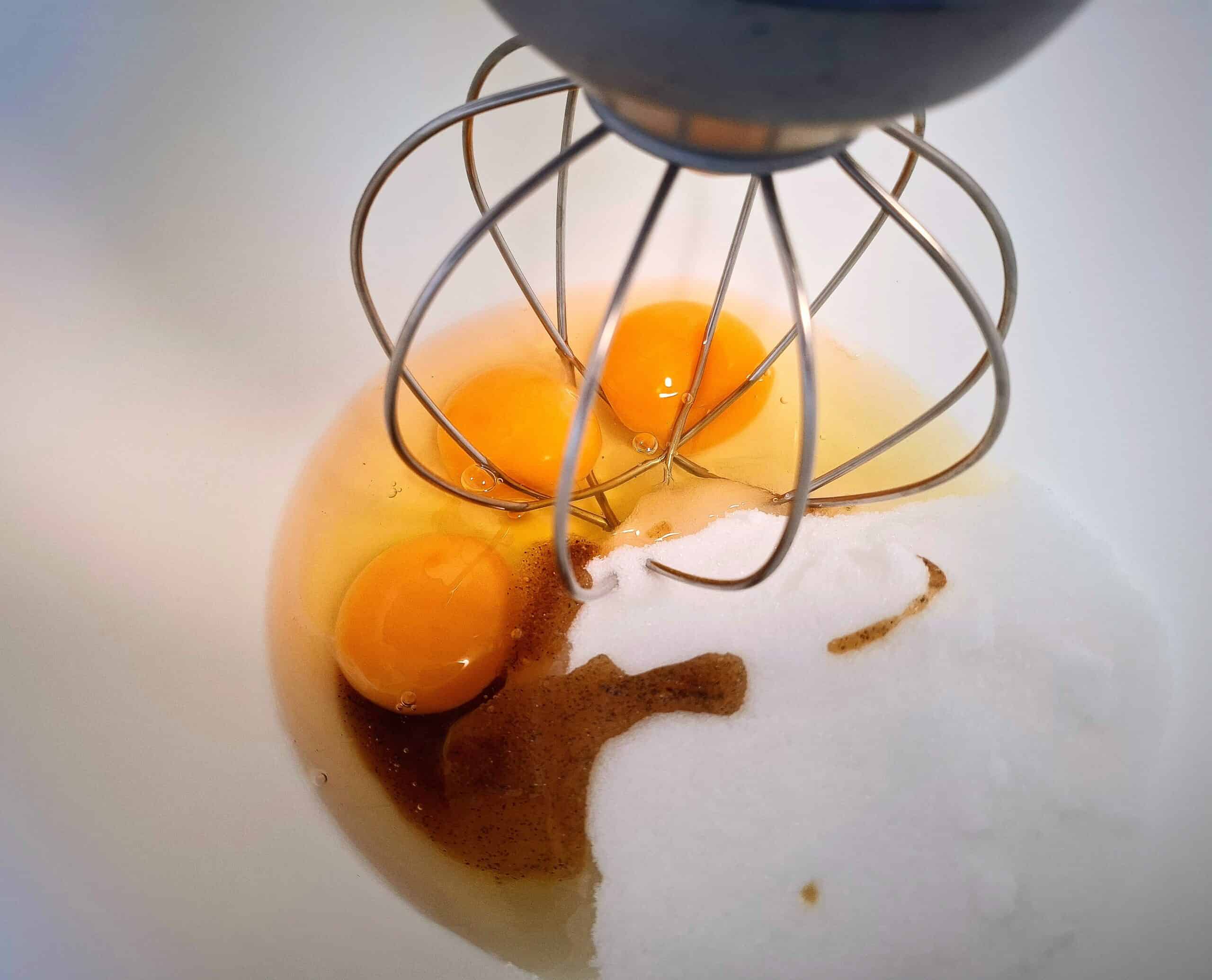 Three cracked eggs, sugar and vanilla paste in a kitchen mixer white bowl and a whisk ready to be mixed.