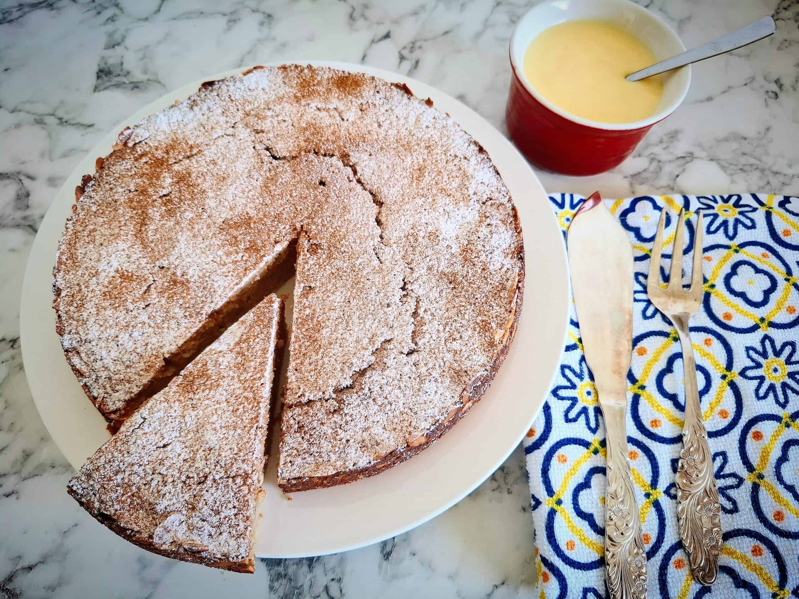 Cooked apple and almond cake dusted with cinnamon and icing sugar on a white round plate on a marbled bench top. A wedge portion has been cut from the cake and the wedge has been pulled away from the rest of the cake slightly. A White, blue and yellow napkin with cake serving cutlery on top next to it and a red ramekin with custard in it above the serving cutlery and next to the cake.