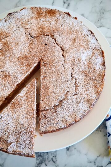 Cooked apple and almond cake dusted with cinnamon and icing sugar on a white round plate on a marbled bench top. A wedge portion has been cut from the cake and the wedge has been pulled away from the rest of the cake slightly. A White, blue and yellow napkin with cake serving cutlery on top next to it.