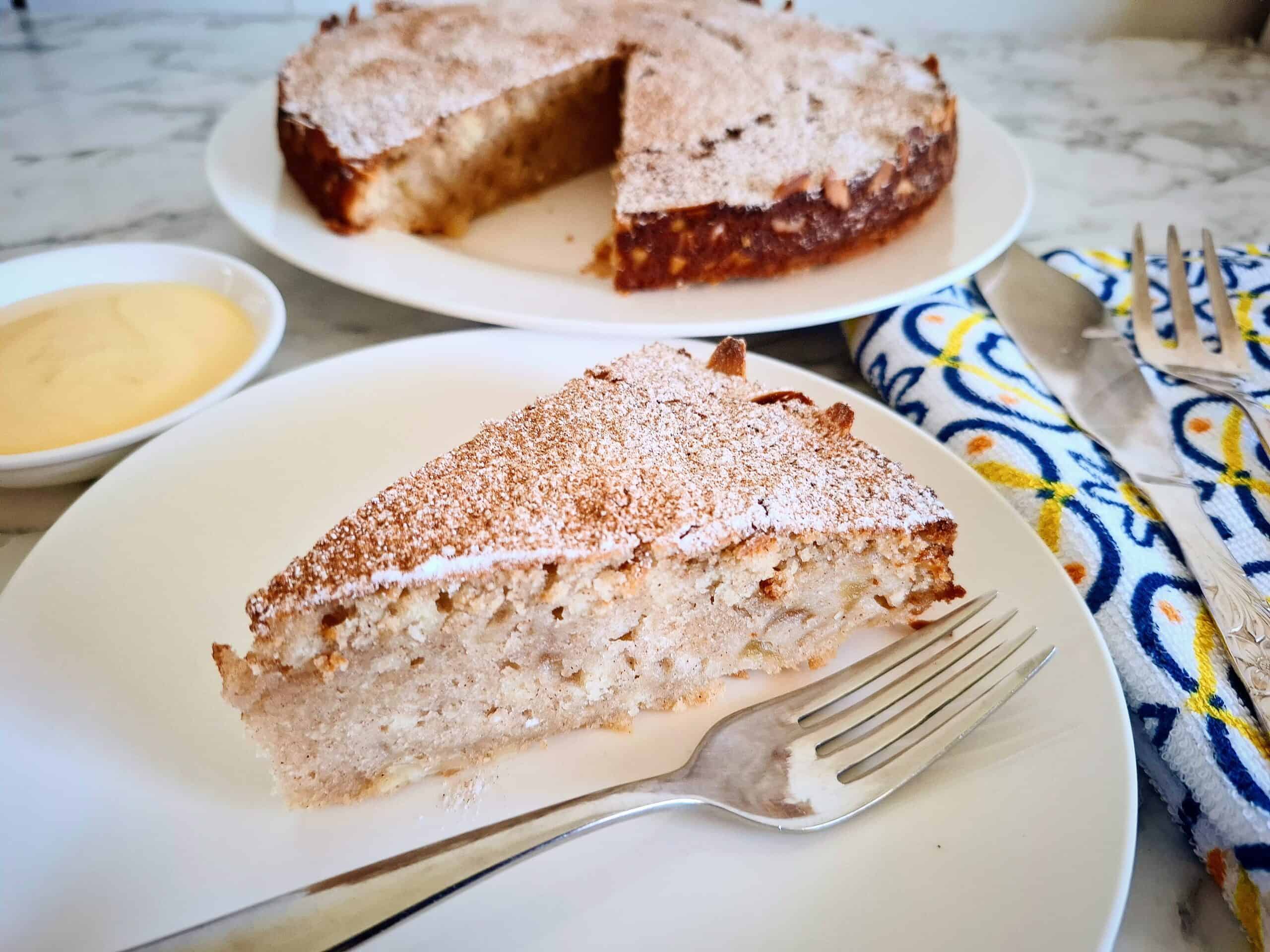 Cooked apple and almond cake dusted with cinnamon and icing sugar on a white round plate on a marbled bench top. A wedge portion has been cut from the cake and served on a white plate with a fork. A White, blue and yellow napkin with cake serving cutlery on top next to it and a white ramekin with custard in it on the other side next to the cake.