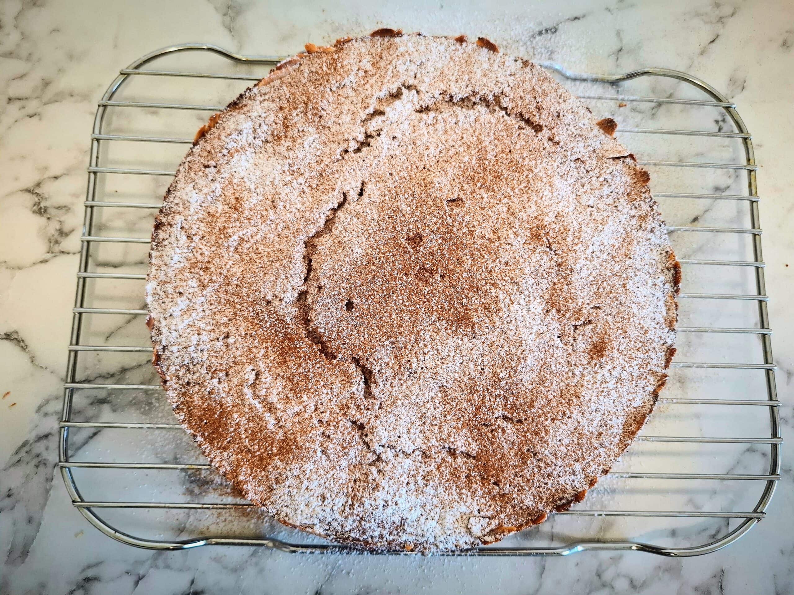 Cooked apple and almond cake dusted with cinnamon and icing sugar on a wired wrack on a marbled bench top.