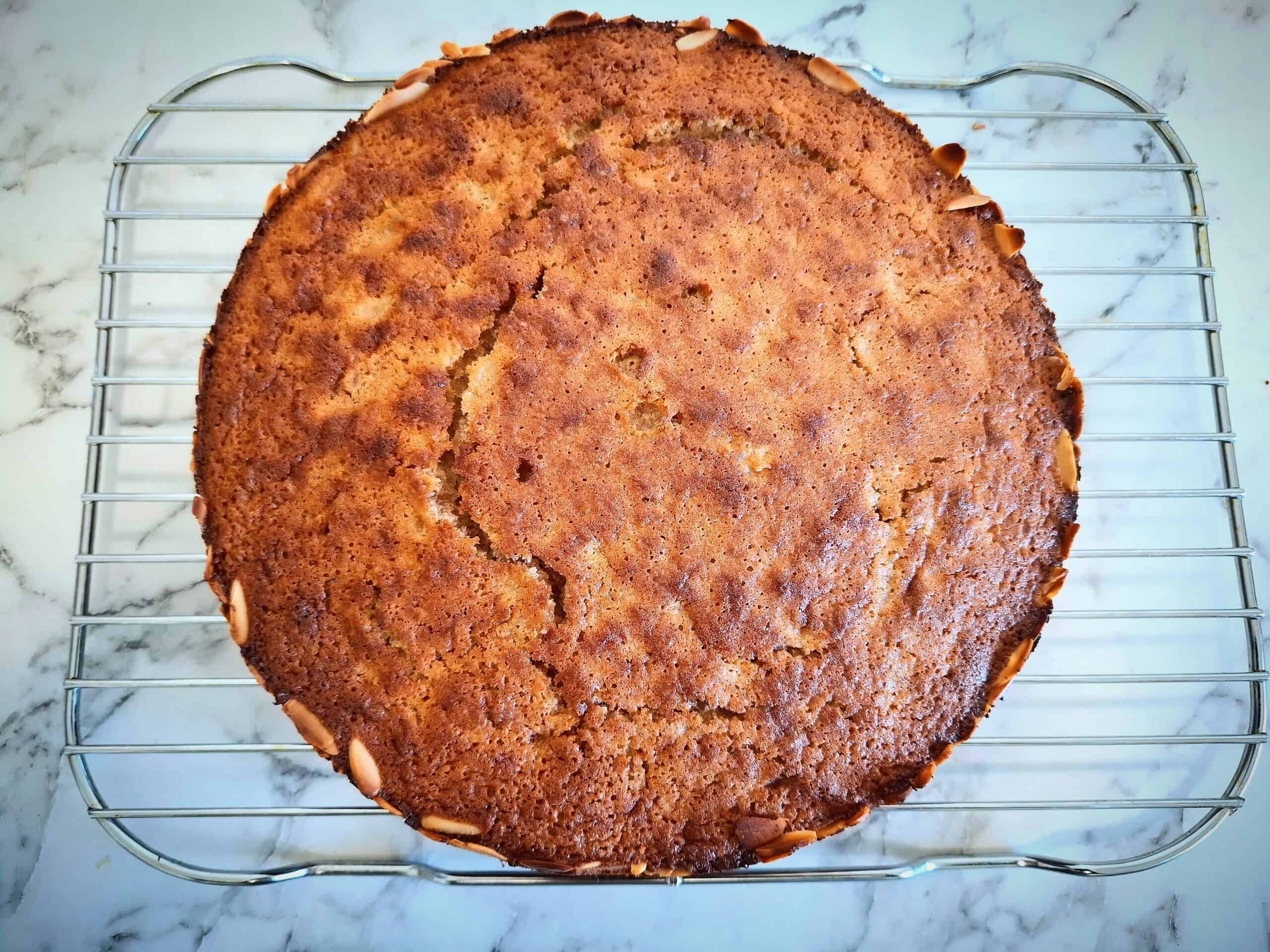 Cooked apple and almond cake cooling down on a wired wrack on a marbled bench top.