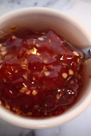 Candied chili in a white ramekin on a marbled bench top. A teaspoon is holding up some candied chili.