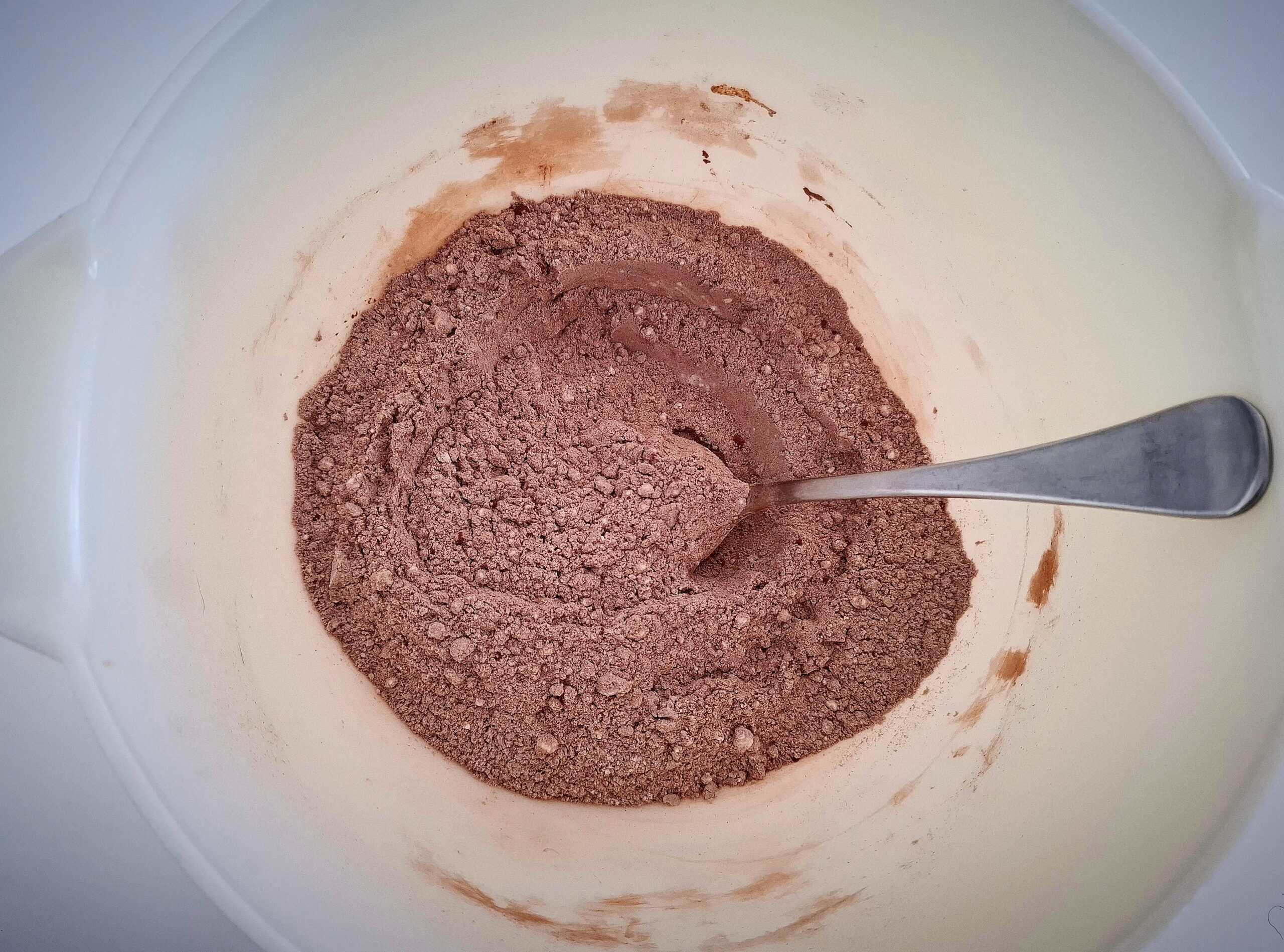 Dry ingredients for chocolate and courgette cake in a white bowl and a silver spoon in it.
