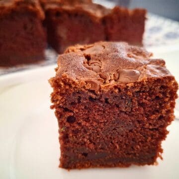 A square piece of courgette and chocolate cake served on a round white plate and a teaspoon on the plate next to the cake. More cut square pieces of the cake served on a blue and white platter behind it.