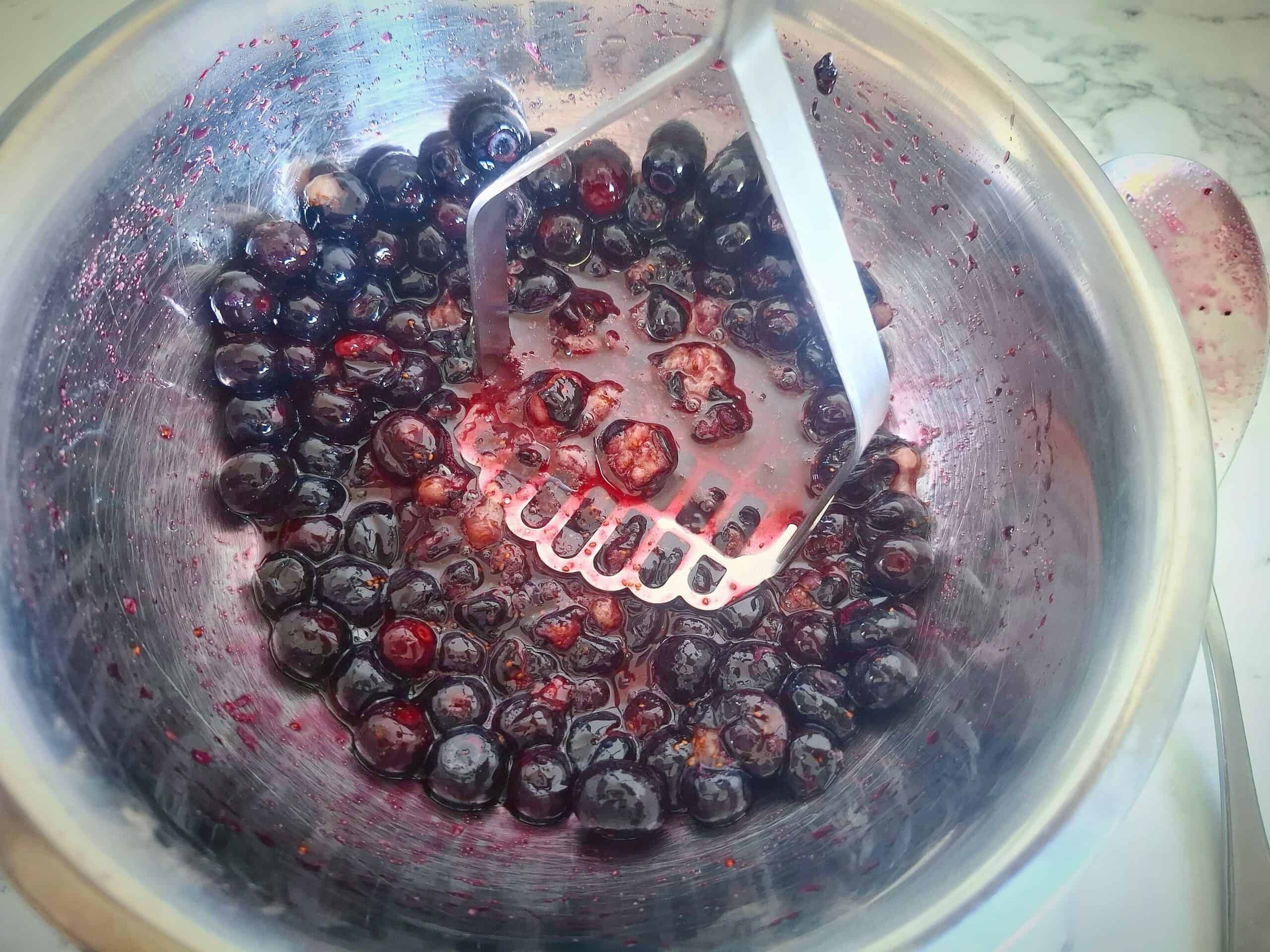 Squashing the blueberries with a potato masher in a stainless steel bowl.