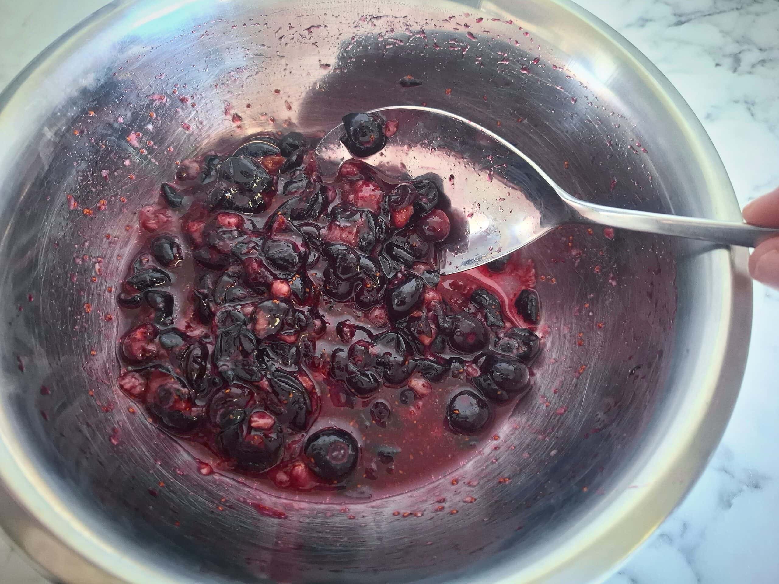 Macerated blueberries in a stainless steel bowl with a silver spoon.