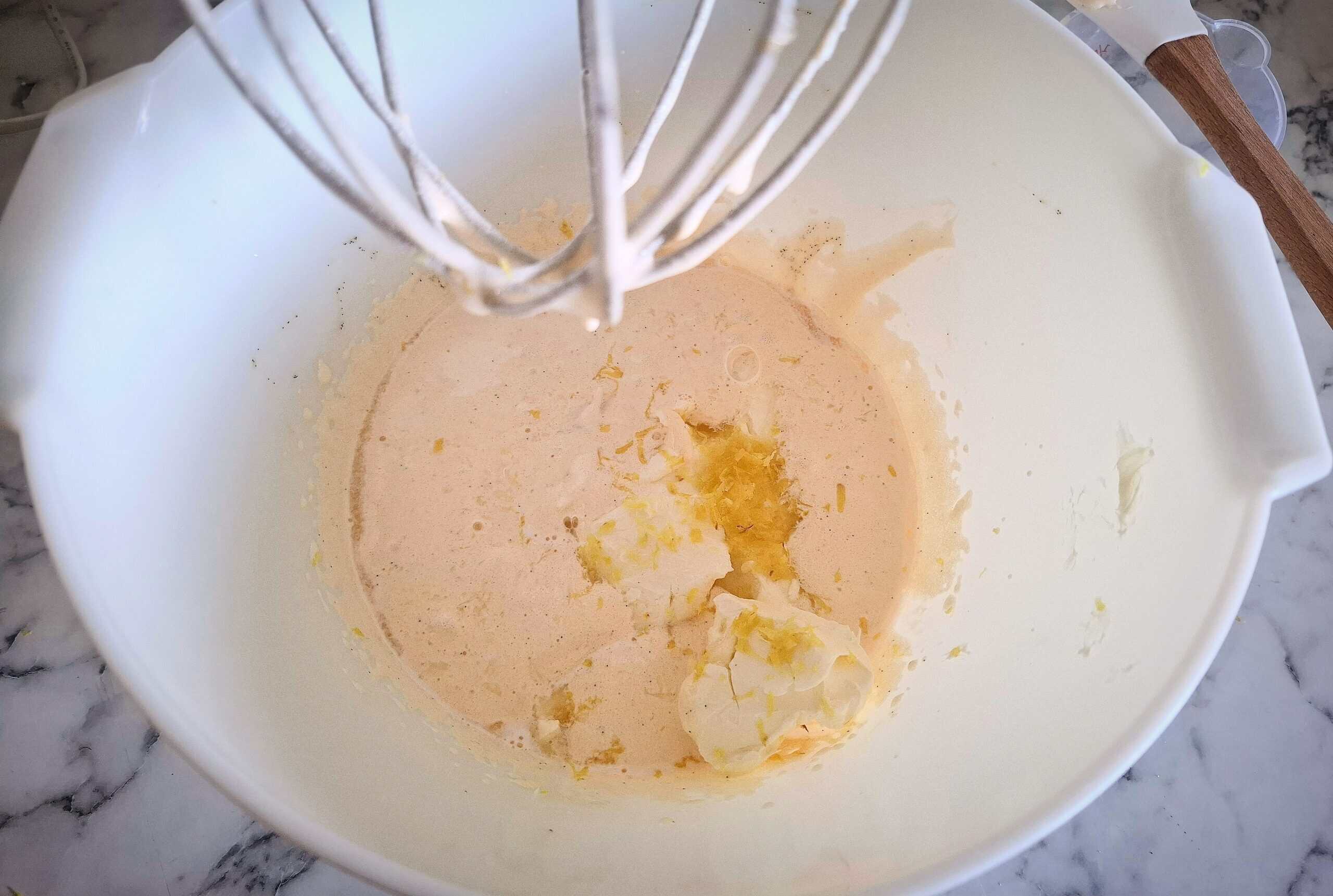 Egg yolks, sugar and vanilla has been whisked white and fluffy. Cream cheese, lemon juice and lemon zest has been added to it in a white kitchen mixer bowl and a whisk above it.