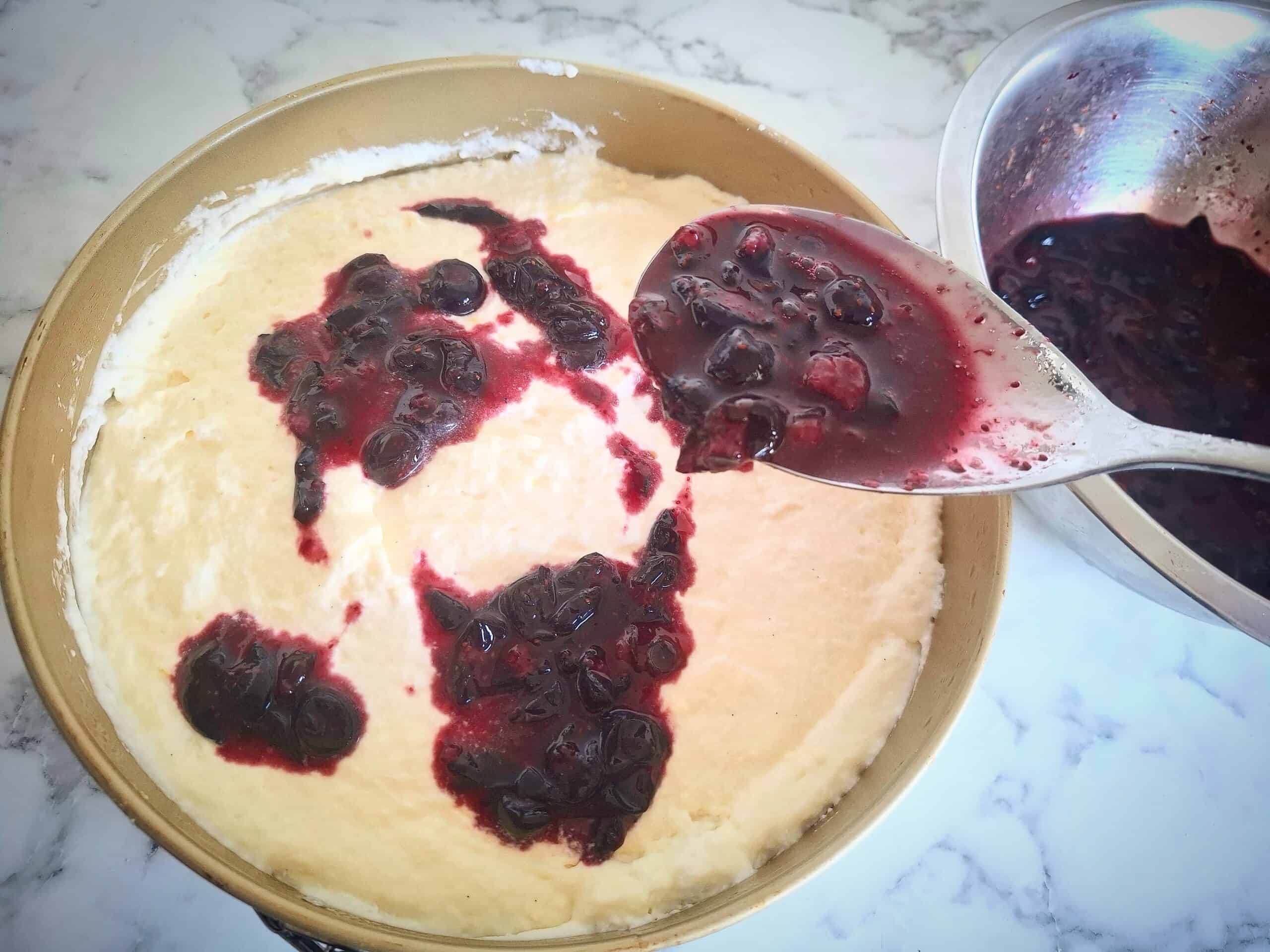 Macerated blueberries drizzled on top of the cheese cake mixture in a round cake tin.