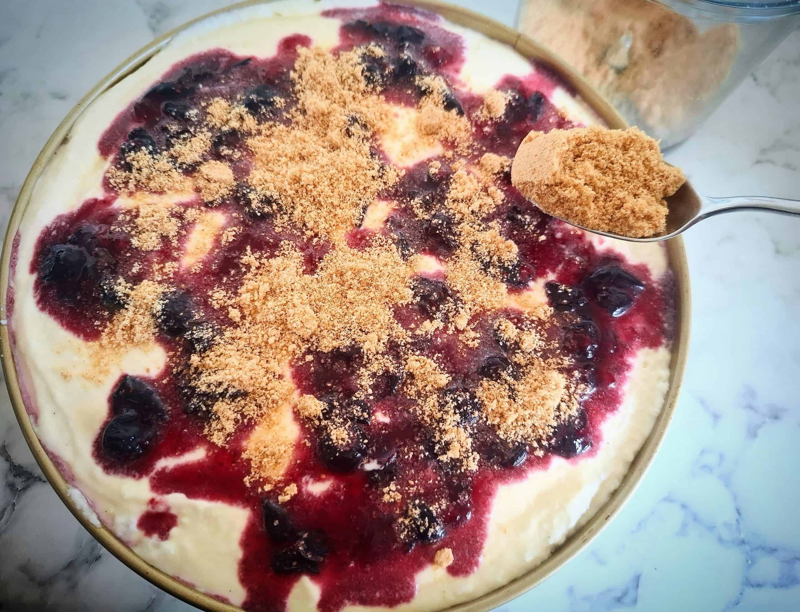 A round cake tin filled with cheese cake mixture, topped with macerated blueberries and digestive biscuit crumbs sprinkled on top.