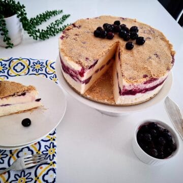 Blueberry frozen cheesecake topped with fresh blueberries on a serving plate. One portion cut out from the main cheesecake and served on a plate next to it on the left hand side, on top of a blue, yellow and white tea towel with a green pot plant behind it. A small ramekin of fresh blueberries and cake serving cutlery in front of the cake on the right hand side.