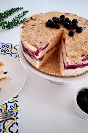 Blueberry frozen cheesecake topped with fresh blueberries on a serving plate. One portion cut out from the main cheesecake and served on a plate next to it on the left hand side, on top of a blue, yellow and white tea towel with a green pot plant behind it. A small ramekin of fresh blueberries and cake serving cutlery in front of the cake on the right hand side.