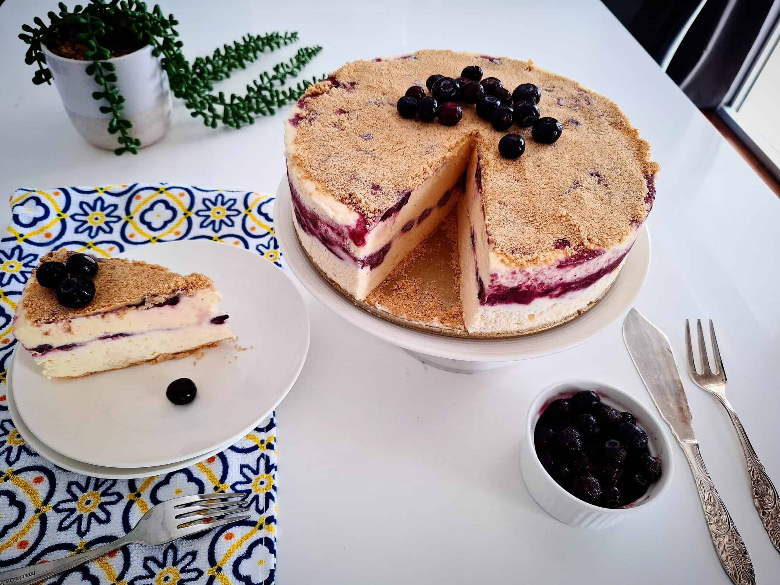 Blueberry frozen cheesecake topped with fresh blueberries on a serving plate. One portion cut out from the main cheesecake and served on a plate next to it on the left hand side, on top of a blue, yellow and white tea towel with a green pot plant behind it. A small ramekin of fresh blueberries and cake serving cutlery in front of the cake on the right hand side.