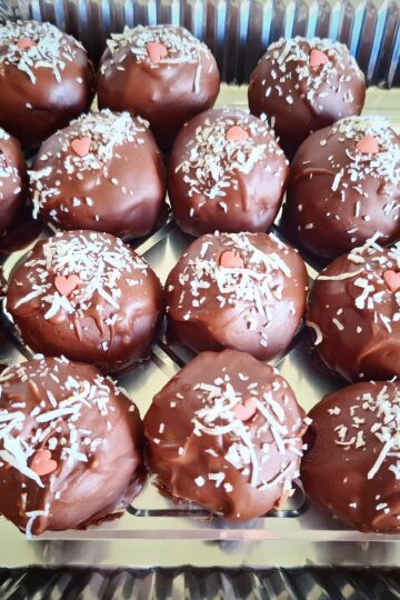 Eighteen chocolate truffle cake ball decorated with shredded coconut and a small pink sugar heart in a foil take away container.