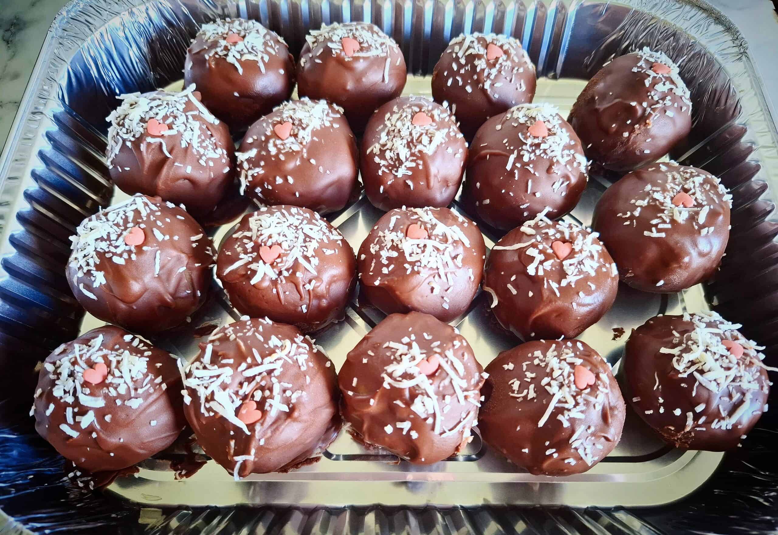 Eighteen chocolate truffle cake ball decorated with shredded coconut and a small pink sugar heart in a foil take away container.
