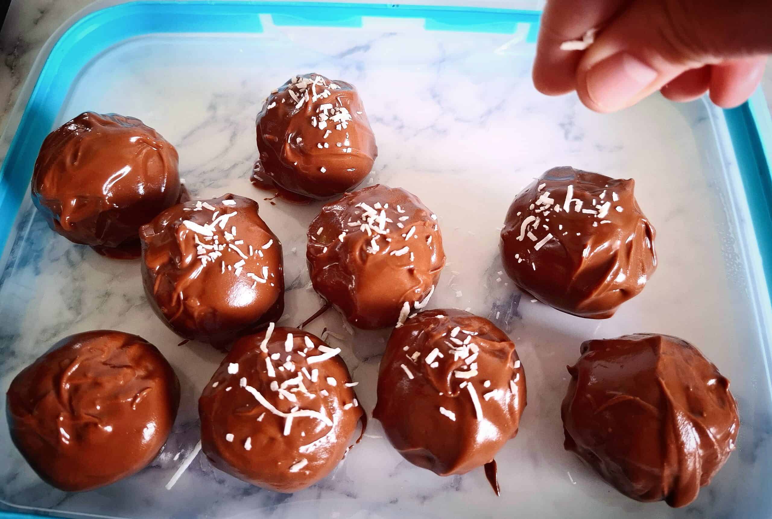 Nine chocolate cake ball truffles on a clear plastic lid with a blue rim. A hand is sprinkling shredded coconut from above