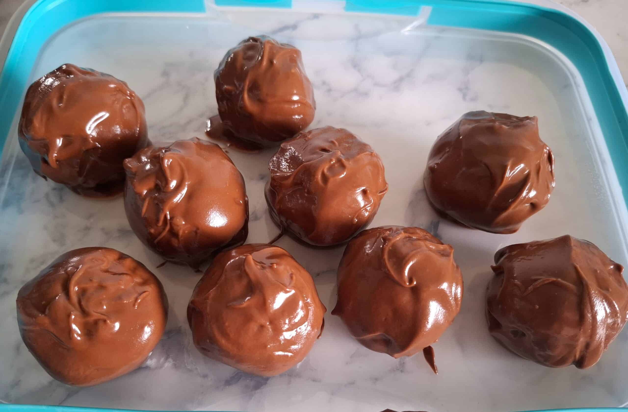 Nine chocolate cake ball truffles freshly coated with melted chocolate on a clear plastic lid with a blue rim.