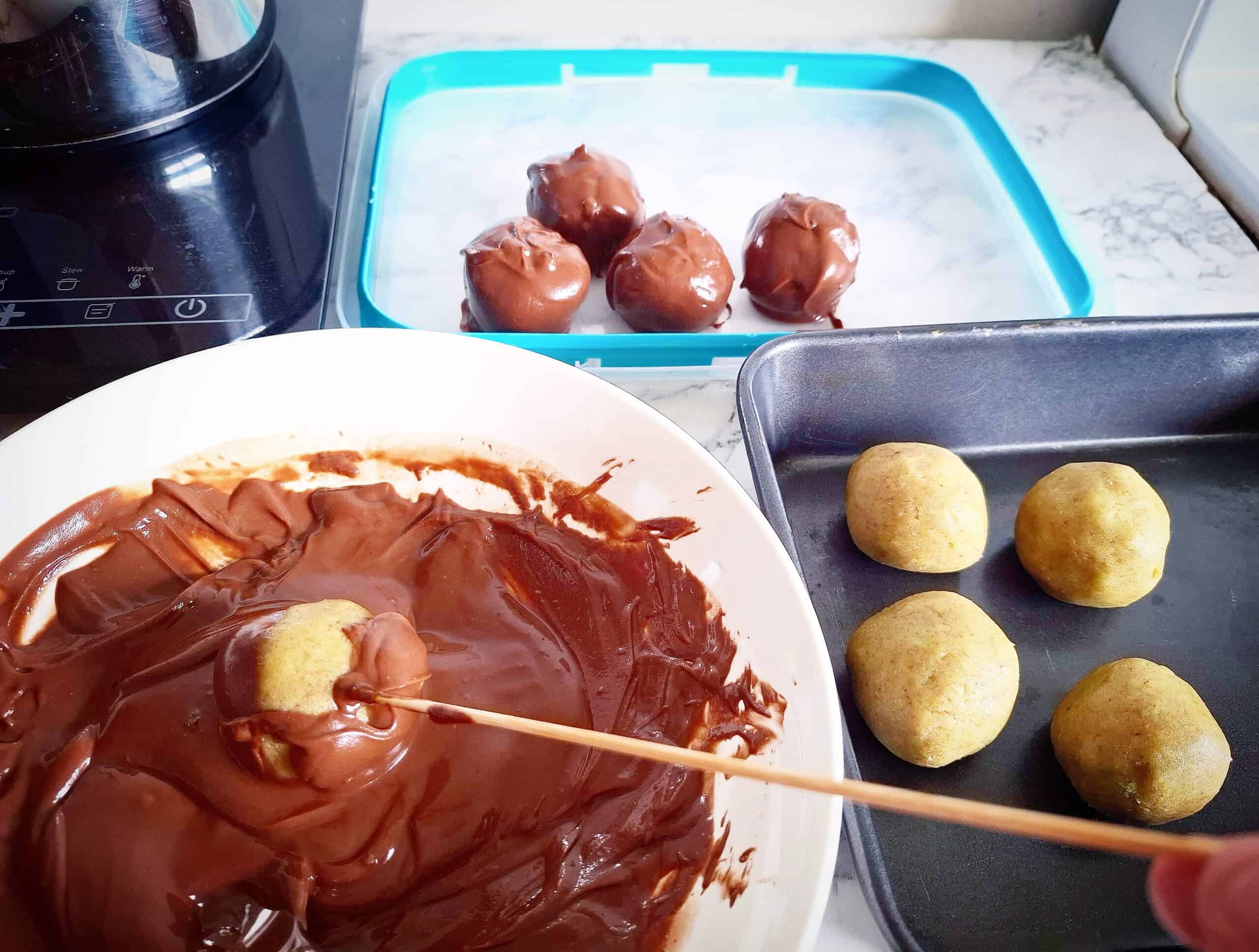 Chocolate cake ball truffles being coated with melted chocolate with the help of a wooden skewer. Four un coated cake balls in a black sheet pan, a white bowl of melted chocolate and a cake ball being coated in it next to the sheet pan. A clear plastic lid with a blue rim behind it with four freshly coated cake truffles on it.