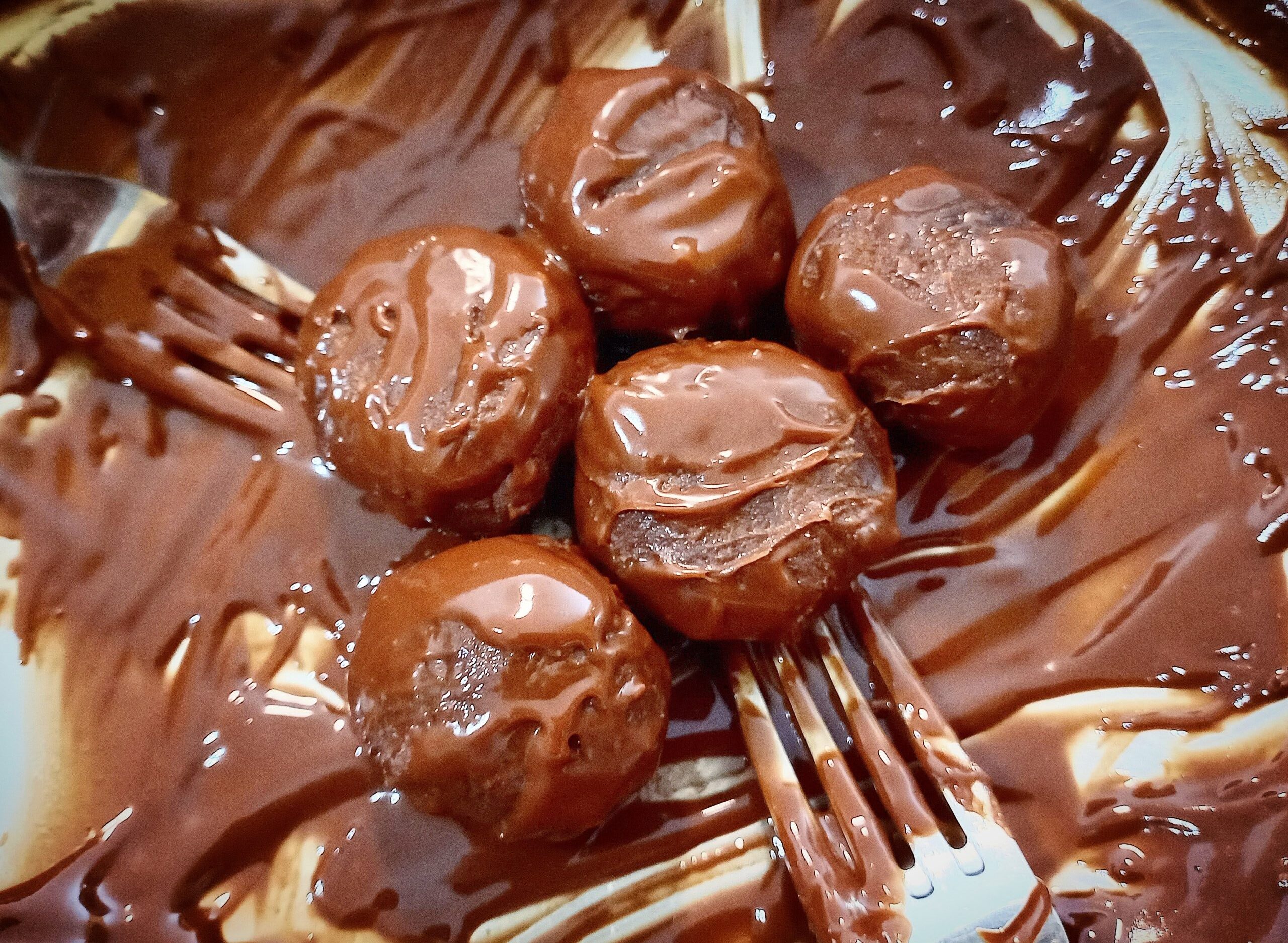 Five chocolate cake ball truffles being coated with melted chocolate with the help of two forks.