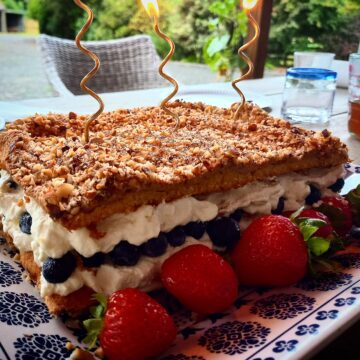 Swedish meringue cake filled with cream and fresh blueberries, decorated with fresh strawberries around it on a blue and white platter. Three cold colored swirl candles in the cake.