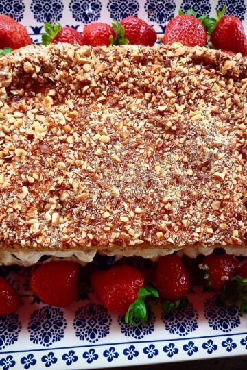 Swedish meringue cake decorated with fresh strawberries around it on a blue and white square platter and a cake serving utensil next to the cake on the platter.