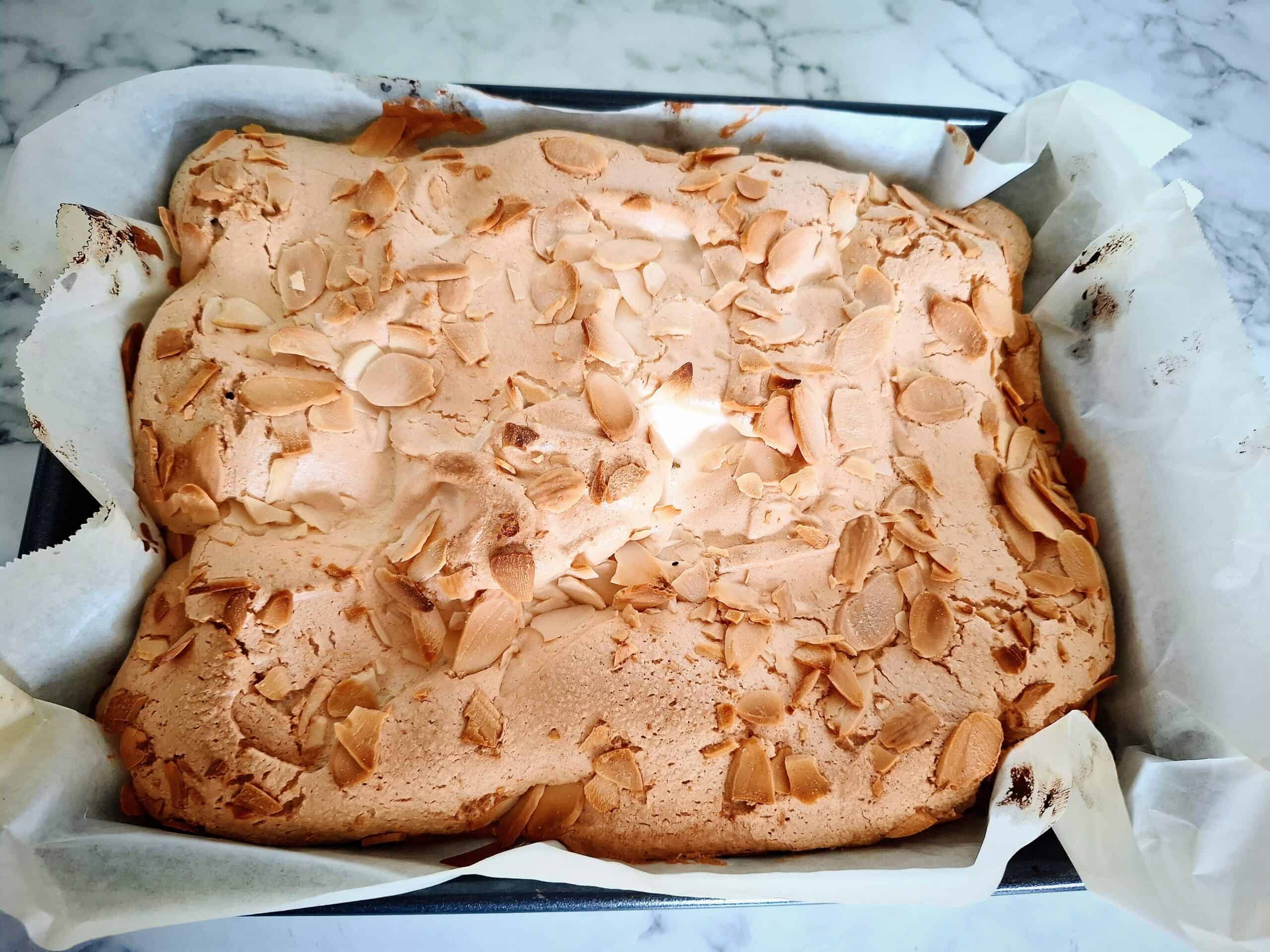 A swedish meringue cake has been cooked in a black rectangular baking tin lined with parchment paper on a marbled bench top.