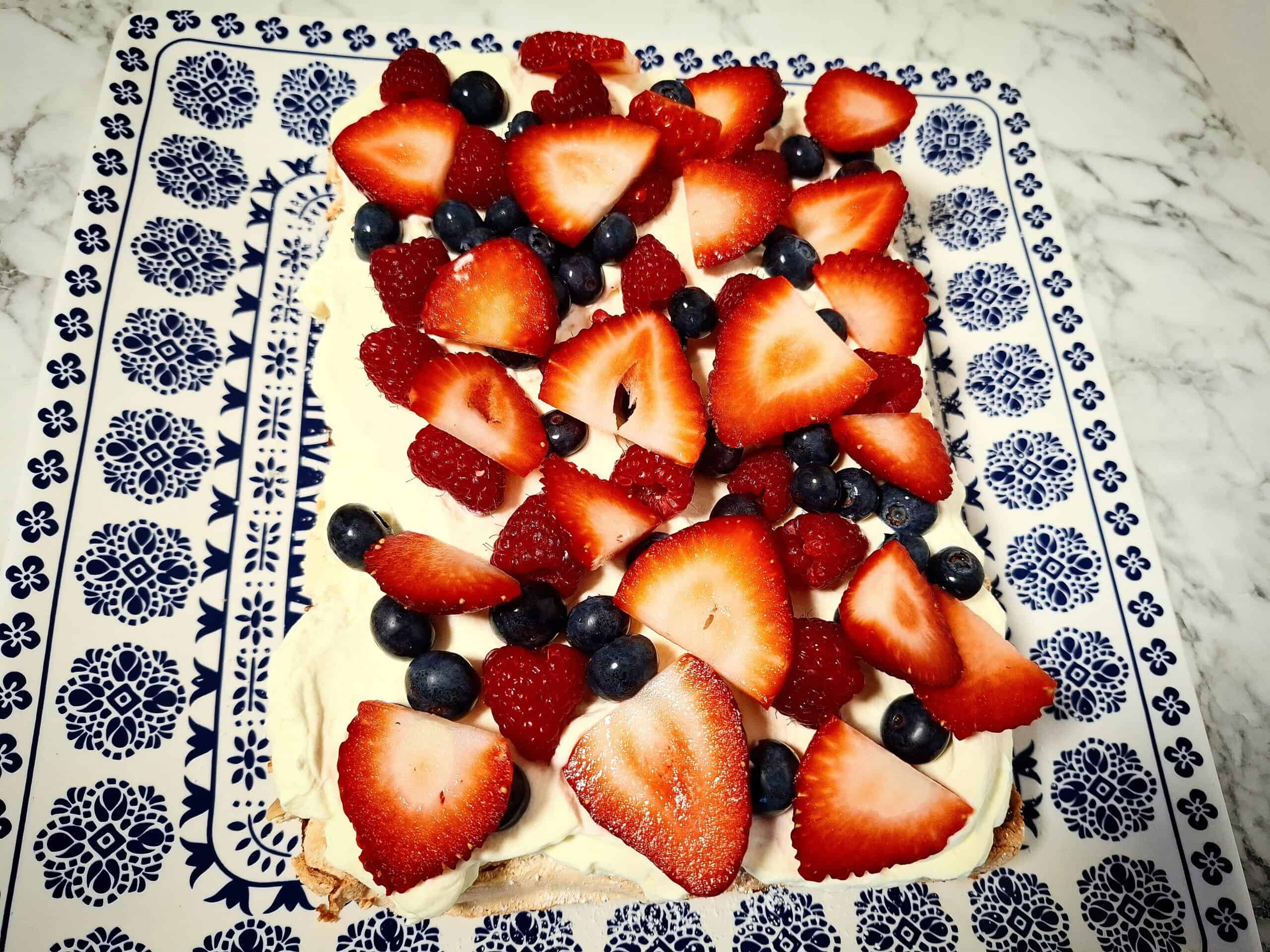 A mixture of fresh berries has been laid out on top of whipped cream on top of a Swedish meringue cake base on a blue and white square platter on a marbled bench top.