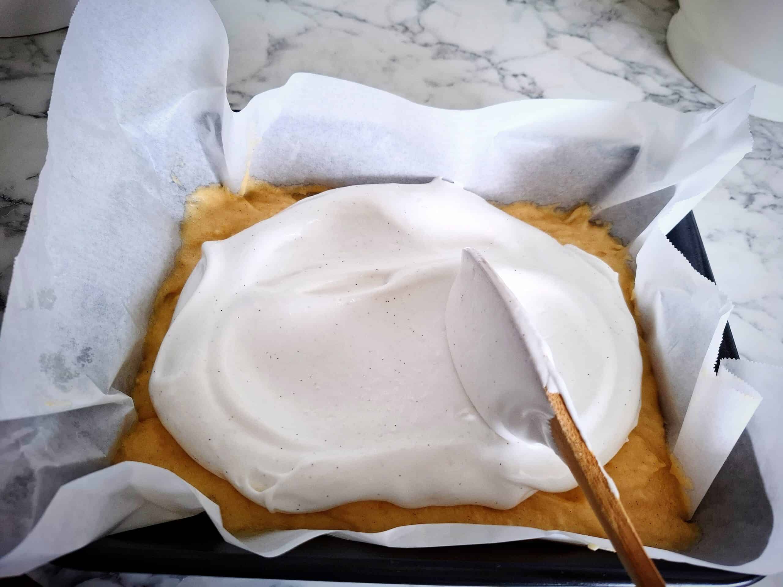Whipped egg whites are being spread on top cake batter in a black rectangular baking tin lined with parchment paper on a marbled bench top.