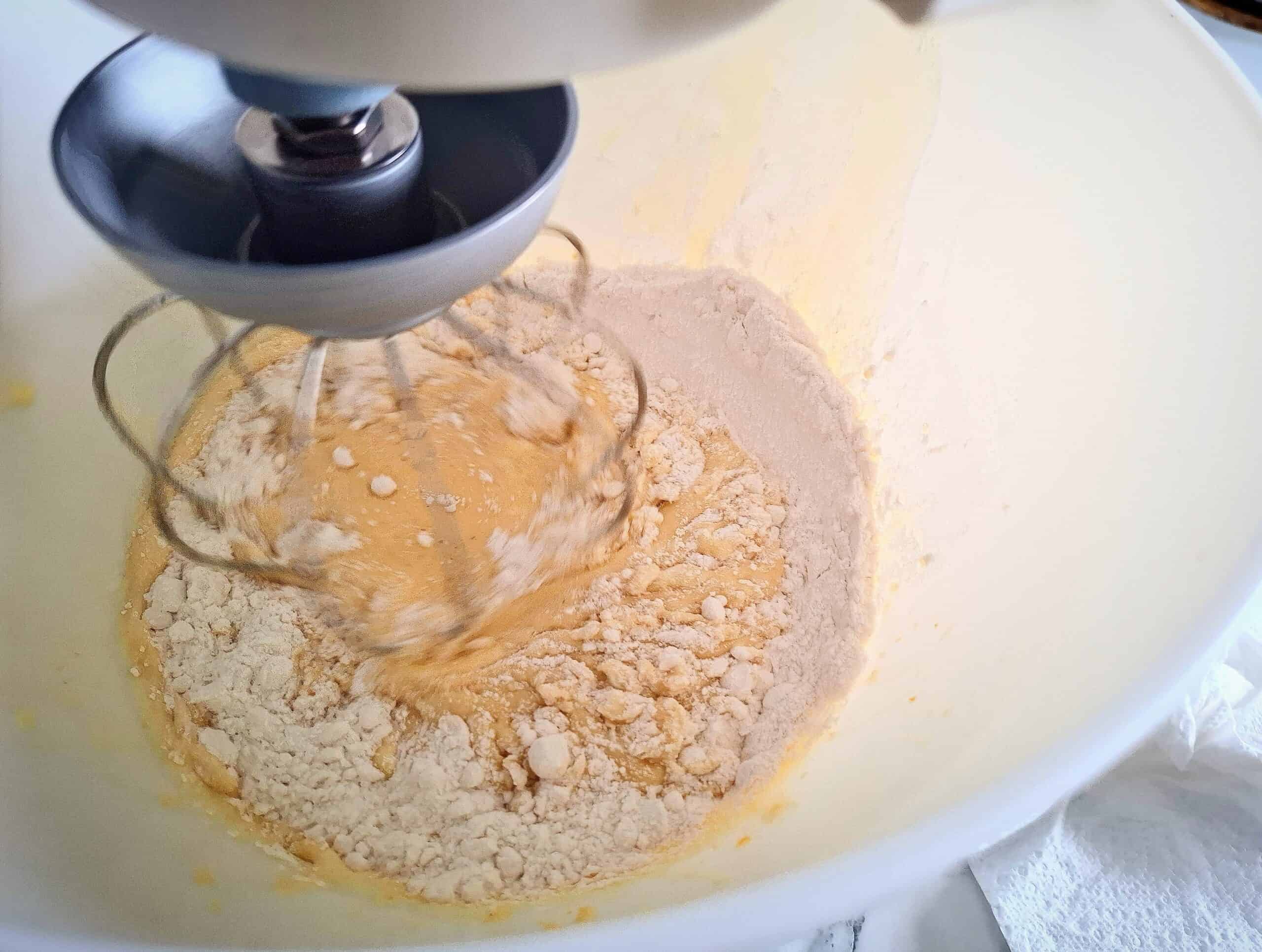 Adding flour to the egg sugar and butter mixture in a white kitchen mixer bowl.