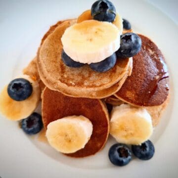 Banana pikelets piled up on a white round plate with fresh blueberries and banana slice on top.